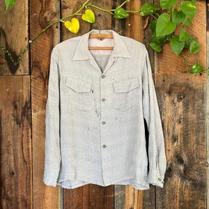 Puede incluir: Una camisa gris claro de manga larga con dos bolsillos en el pecho y cuello. La camisa tiene una botonadura frontal y puños abotonados. La tela tiene un aspecto texturizado. La camisa está colgada en una percha de madera sobre un fondo de madera.