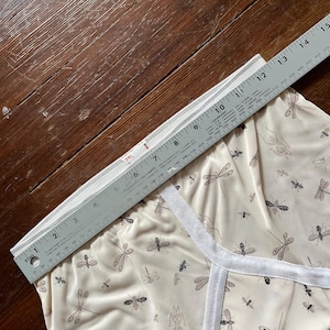 Op de afbeelding: Cr&egrave;mekleurige pyjamashorts met een libellenpatroon, gemeten met een liniaal. De shorts hebben een witte bies en zijn gemaakt van een zachte stof. De liniaal toont een afmeting van ongeveer 38 cm.