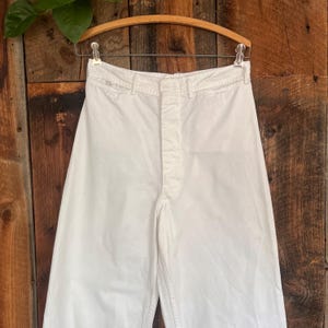 May include: High-waisted white trousers featuring a button-fly closure, a structured waistband with belt loops, and a clean, solid-colored design. The pants are displayed on a wooden hanger, showcasing a straight-leg cut and a crisp, plain finish.