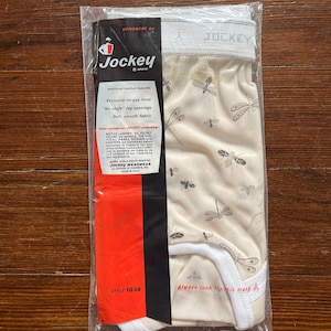 Op de afbeelding: Verzegeld pakket Jockey-ondergoed. Het ondergoed is beige met een libellenpatroon. De verpakking bevat het Jockey-logo en de tekst "Exclusive no-gap front" en "Soft, smooth fabric."