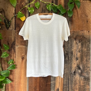 Puede incluir: Una camiseta crema de manga corta colgada de una percha de madera. La camiseta tiene cuello redondo y un diseño sencillo y clásico. El fondo es una pared de madera rústica con follaje verde.