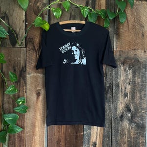 Könnte beinhalten: Schwarzes T-Shirt mit einer weißen Grafik von Tommy Bolin, einem Musiker, mit dem Text "Tommy Bolin" in weiß.