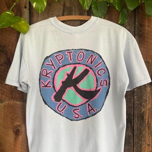 May include: Light blue t-shirt with a graphic of a circle with the text "Kryptonics U.S.A." in a blue, pink, and green color scheme.