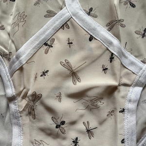 Op de afbeelding: Close-up van beige ondergoed met een herhaald patroon van libellen en andere insecten in bruin- en zwarttinten. De randen zijn afgewerkt met witte stof.