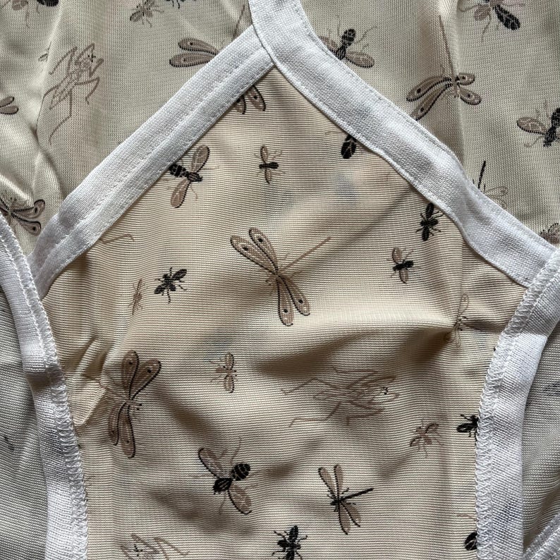 Op de afbeelding: Close-up van beige ondergoed met een herhaald patroon van libellen en andere insecten in bruin- en zwarttinten. De randen zijn afgewerkt met witte stof.