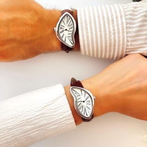 May include: Two wristwatches with a unique, melting design. The watch face is white with black Roman numerals and hands, set in a silver-tone case. The brown leather straps are attached to the watch case. The watches are worn on wrists.