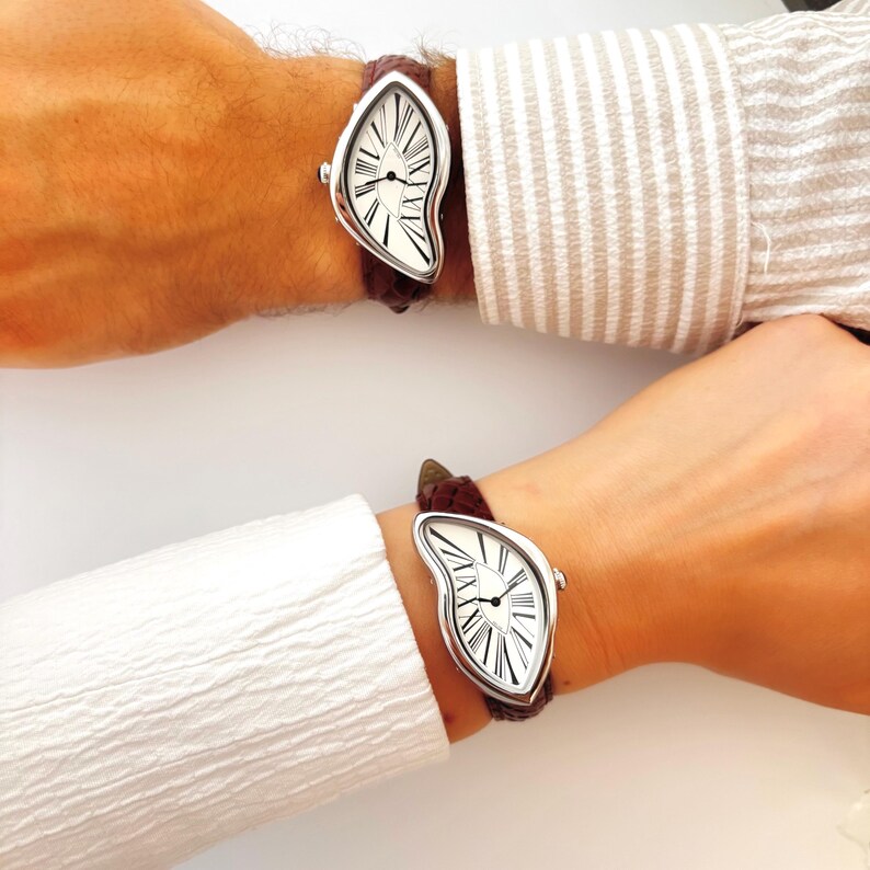 May include: Two wristwatches with a unique, melting design. The watch face is white with black Roman numerals and hands, set in a silver-tone case. The brown leather straps are attached to the watch case. The watches are worn on wrists.