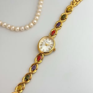May include: Gold-toned wristwatch with a white face and Roman numerals. The watch band features colorful oval-shaped gemstones. A strand of pearls is in the background. The watch is marked "FELISE".