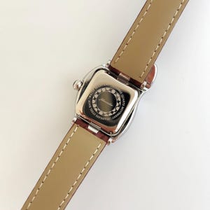 May include: A silver-toned wristwatch with a brown leather band. The watch face is square with rounded edges. The back of the watch has the text "Stainless Steel Back, Water Resistant, Japanese Quartz Movement" and the brand name "Johnwacher".