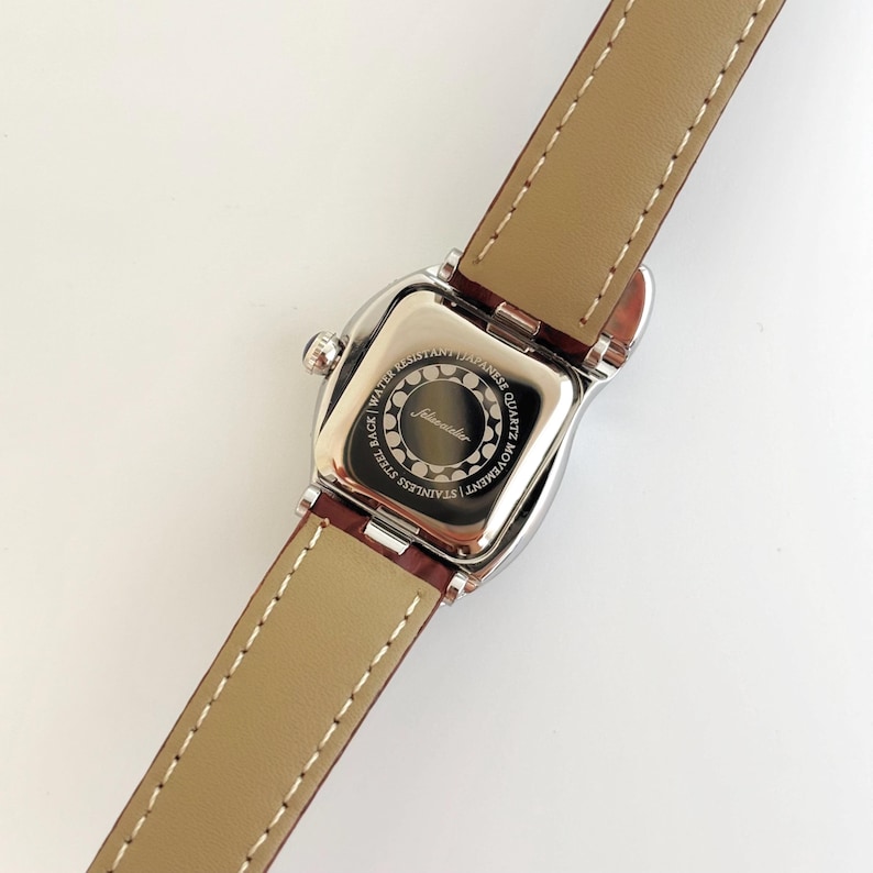 May include: A silver-toned wristwatch with a brown leather band. The watch face is square with rounded edges. The back of the watch has the text "Stainless Steel Back, Water Resistant, Japanese Quartz Movement" and the brand name "Johnwacher".