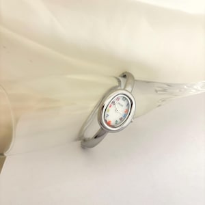 Puede incluir: Reloj de pulsera plateado con esfera ovalada. La esfera del reloj es blanca con marcadores de hora coloridos y agujas negras. La pulsera tiene un dise&ntilde;o simple y curvado. Un accesorio elegante.