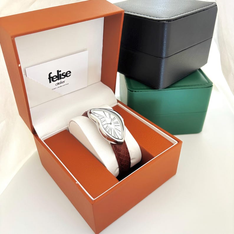 May include: A silver-toned watch with a white face and Roman numerals, featuring a unique, melting design. The watch has a brown leather band and is displayed in an orange box with the brand name "felise atelier". Additional black and green boxes are stacked in the background.