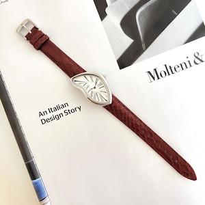 May include: A silver-toned wristwatch with a white face and black Roman numerals, featuring a unique, melting design. The watch has a dark red, textured leather band. The text "An Italian Design Story" is visible.