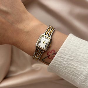 May include: A rectangular wristwatch featuring a white dial with black Roman numerals and gold-tone hands. The watch case and the multi-link bracelet are constructed from a combination of silver-tone and gold-tone metal. The brand name FELISE is printed on the dial.