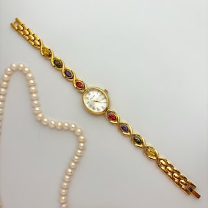 May include: A gold-toned wristwatch with an oval face and Roman numerals. The watch band features colorful gemstones. A strand of pearls is in the background. The watch is a Celine brand.