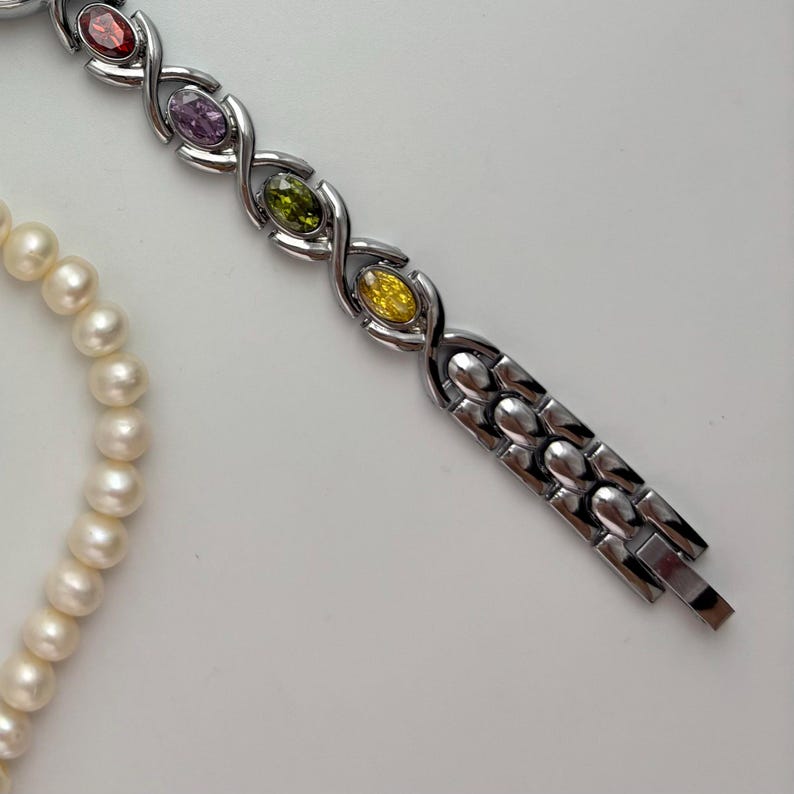 May include: A silver-toned bracelet with oval-shaped, colorful gemstones in red, purple, green, and yellow. A strand of white pearls is also visible, creating a contrast against the bracelet's design. The bracelet has a clasp closure.