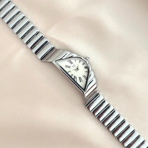 Op de afbeelding: Een zilverkleurig polshorloge met een witte wijzerplaat en zwarte Romeinse cijfers. Het horloge heeft een uniek, asymmetrisch ontwerp met een flexibele band. De wijzerplaat is ongeveer 2,5 cm lang.