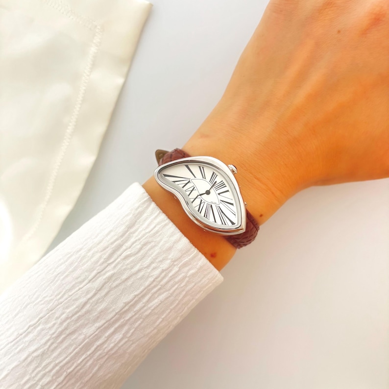 May include: A silver-toned wristwatch with a melting clock face design and Roman numeral hour markers. The watch has a brown leather band. The watch face is white with black hands and hour markers. The watch is a unique and artistic accessory.