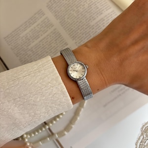 May include: A silver wristwatch with a woven strap and a round, white face. The watch features Roman numeral hour markers and a textured bezel. The watch is worn on a wrist. The design is elegant and classic. The strap is approximately 15 cm in length.