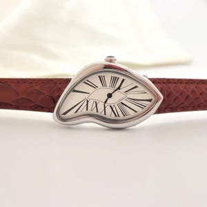 May include: A silver-toned wristwatch with a unique, melting design. The watch face is white with black Roman numerals and hands. It has a brown leather band. The watch has a small blue gem on top.