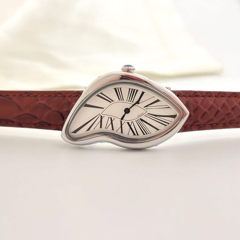 May include: A silver-toned wristwatch with a unique, melting design. The watch face is white with black Roman numerals and hands. It has a brown leather band. The watch has a small blue gem on top.