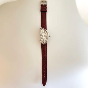 May include: A silver-toned wristwatch with a melting clock face design, featuring black Roman numerals on a white background. The watch is attached to a burgundy leather strap with a silver buckle. The strap has a textured, snake-like pattern.