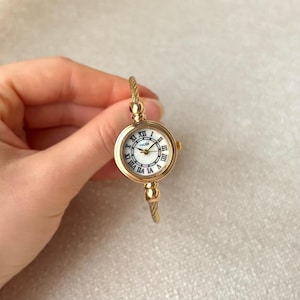 Op de afbeelding: Goudkleurig polshorloge met een witte wijzerplaat en zwarte Romeinse cijfers. Het horloge heeft een ronde wijzerplaat en een goudkleurige, gedraaide band. Het horloge wordt in een hand gehouden.
