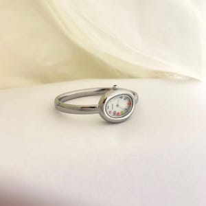 Puede incluir: Un reloj de anillo plateado con una esfera ovalada. La esfera del reloj es blanca con n&uacute;meros negros y detalles coloridos. La banda es un dise&ntilde;o simple de metal pulido.
