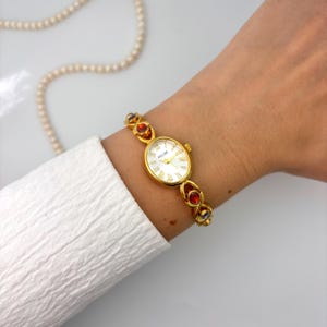 May include: Gold-toned Felise watch with a white face and Roman numerals. The watch band features red, orange, and blue gemstones. A white shirt sleeve and a pearl necklace are in the background.
