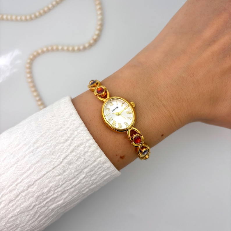 May include: Gold-toned Felise watch with a white face and Roman numerals. The watch band features red, orange, and blue gemstones. A white shirt sleeve and a pearl necklace are in the background.