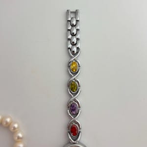 May include: Silver-toned wristwatch with a band featuring yellow, green, purple, and red oval-shaped gemstones. The watch is set against a white background, with a pearl necklace partially visible on the left.