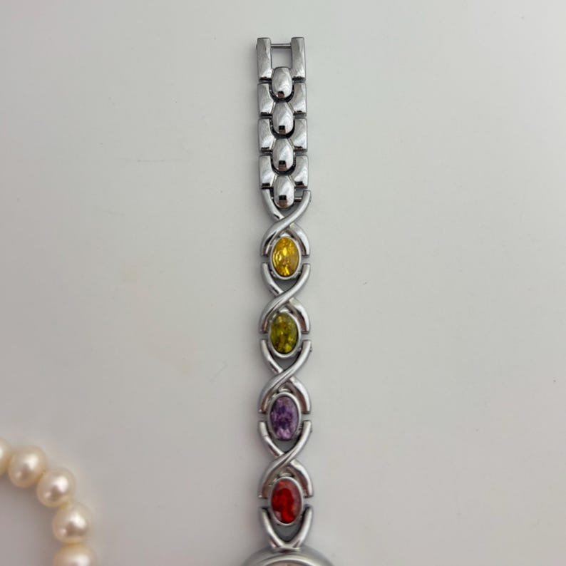 May include: Silver-toned wristwatch with a band featuring yellow, green, purple, and red oval-shaped gemstones. The watch is set against a white background, with a pearl necklace partially visible on the left.