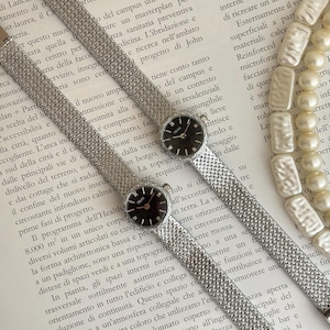 Op de afbeelding: Twee zilverkleurige polshorloges met mesh bandjes en zwarte wijzerplaten. De horloges zijn diagonaal geplaatst en tonen een klassiek ontwerp. Een parelketting is zichtbaar op de achtergrond.