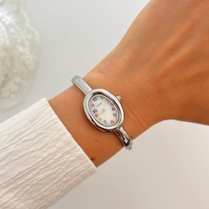 Puede incluir: Reloj ovalado plateado con esfera blanca y n&uacute;meros coloridos. El reloj tiene una correa plateada y se lleva en la mu&ntilde;eca. La esfera del reloj tiene la palabra "FELISE" impresa.