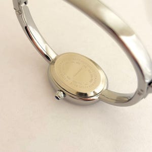 Puede incluir: Un reloj de pulsera plateado con una esfera ovalada. El reloj tiene un movimiento de cuarzo y una parte trasera de acero inoxidable. El reloj tiene una peque&ntilde;a gema azul en el lateral de la esfera.