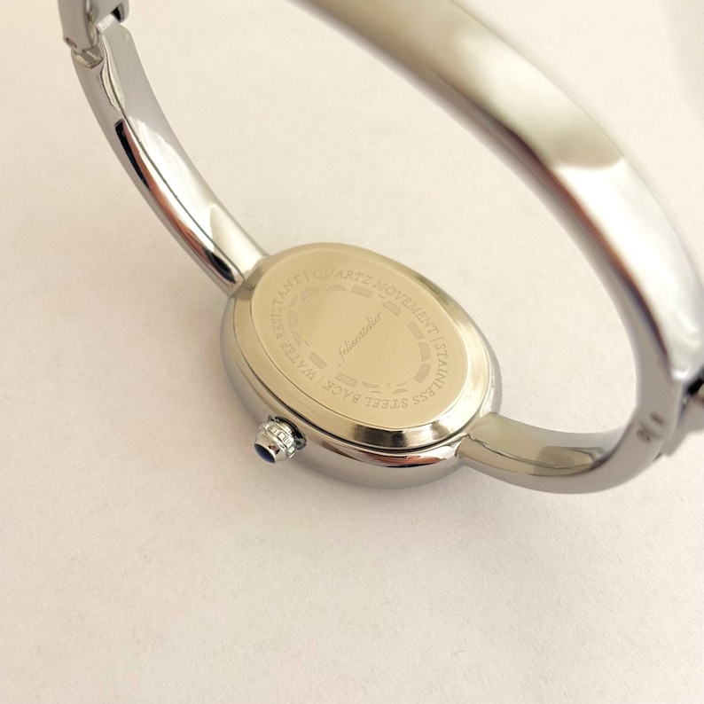 Puede incluir: Un reloj de pulsera plateado con una esfera ovalada. El reloj tiene un movimiento de cuarzo y una parte trasera de acero inoxidable. El reloj tiene una peque&ntilde;a gema azul en el lateral de la esfera.