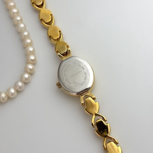 May include: A gold-toned wristwatch with an oval face and a matching bracelet-style band. The watch face is silver with the text "felissandier". A white pearl necklace is partially visible in the image.