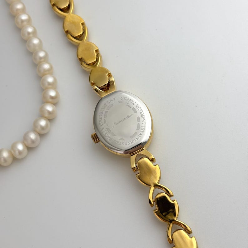 May include: A gold-toned wristwatch with an oval face and a matching bracelet-style band. The watch face is silver with the text "felissandier". A white pearl necklace is partially visible in the image.
