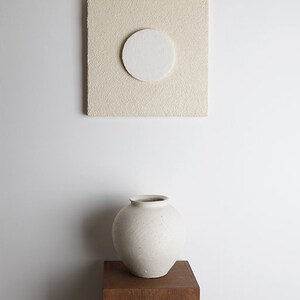 May include: A white ceramic vase with a textured surface sits on a wooden pedestal against a white wall. A square beige canvas with a white circle in the center hangs on the wall above the vase.