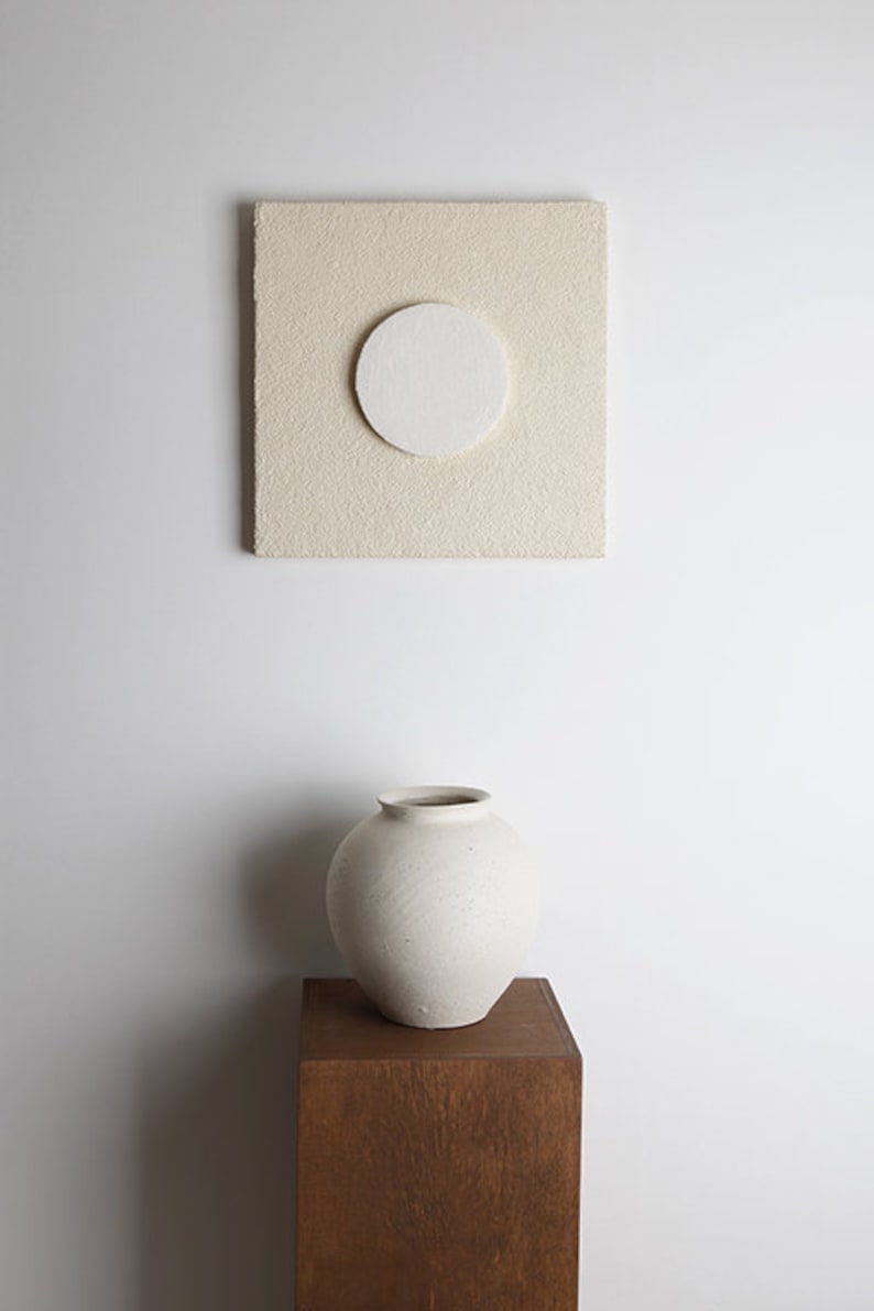 May include: A white ceramic vase with a textured surface sits on a wooden pedestal against a white wall. A square beige canvas with a white circle in the center hangs on the wall above the vase.