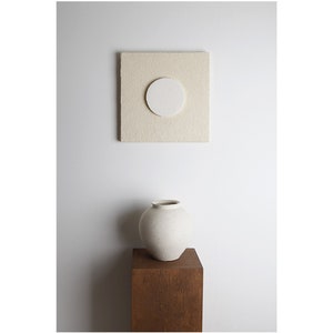 May include: A white ceramic vase with a round body sits on a wooden pedestal in front of a white wall. A square beige canvas with a white circle in the center hangs on the wall.