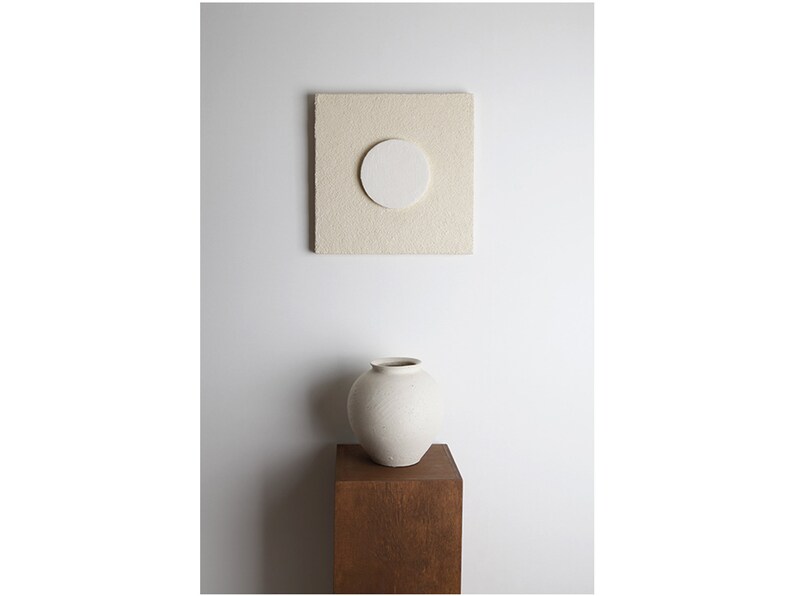 May include: A white ceramic vase with a round body sits on a wooden pedestal in front of a white wall. A square beige canvas with a white circle in the center hangs on the wall.