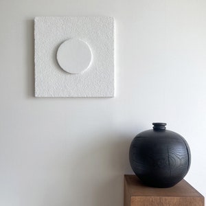 May include: A white textured square canvas with a white circle in the center, hanging on a white wall. Below the canvas is a black wooden vase on a wooden pedestal.