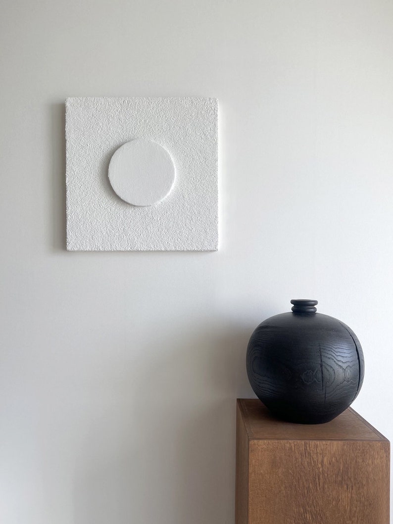 May include: A white textured square canvas with a white circle in the center, hanging on a white wall. Below the canvas is a black wooden vase on a wooden pedestal.