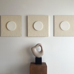 May include: Three off-white square canvases with a circular cutout in the center. Each canvas has a white circle in the cutout. A small beige abstract sculpture sits on a wooden pedestal in front of the canvases.