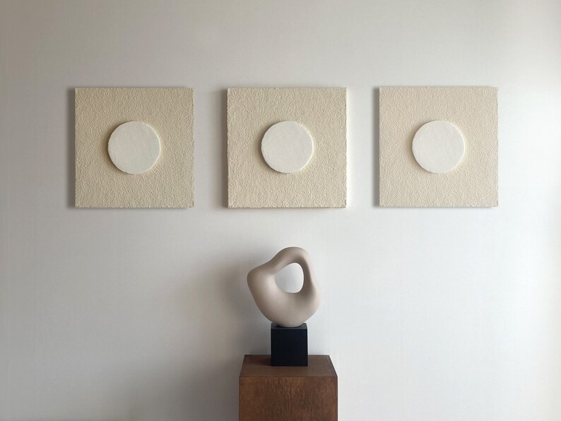 May include: Three off-white square canvases with a circular cutout in the center. Each canvas has a white circle in the cutout. A small beige abstract sculpture sits on a wooden pedestal in front of the canvases.