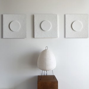 May include: Three white square wall art pieces with a circle cutout in the center. The wall art is hanging on a white wall. A white paper lamp sits on a wooden table in front of the wall art.