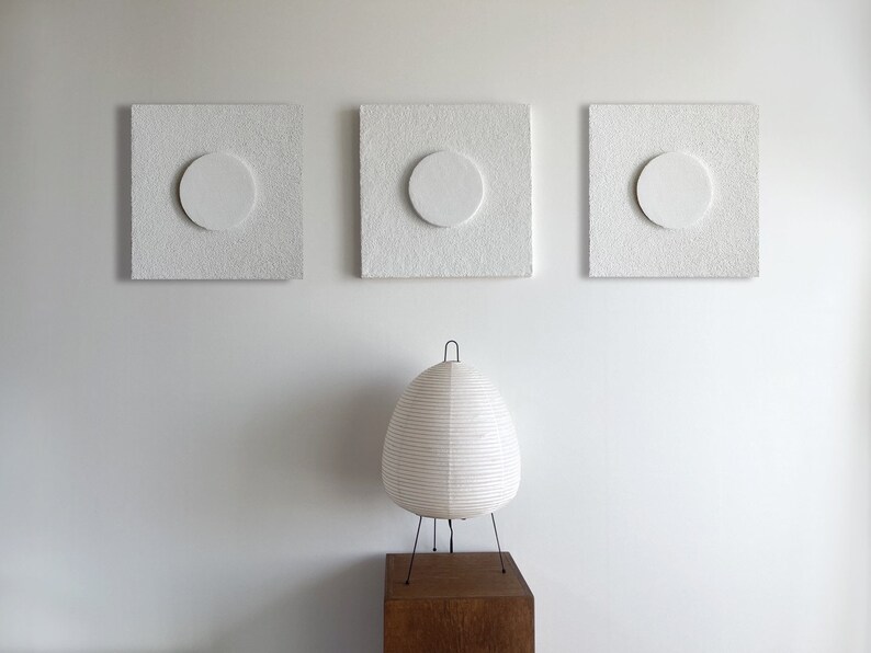 May include: Three white square wall art pieces with a circle cutout in the center. The wall art is hanging on a white wall. A white paper lamp sits on a wooden table in front of the wall art.