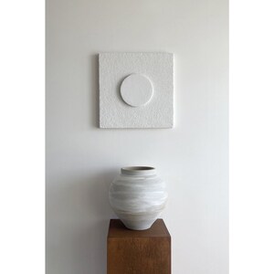 May include: A white ceramic vase with a textured surface sits on a wooden stand in front of a white square canvas with a white circle in the center.