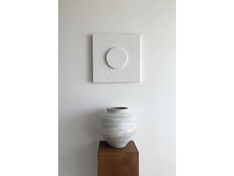 May include: A white ceramic vase with a textured surface sits on a wooden stand in front of a white square canvas with a white circle in the center.